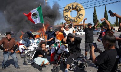Manifestantes en Los Ángeles se congregan en una protesta con vehículos incendiados de fondo. Algunos portan la bandera de México mientras posan frente al fuego, en un acto de desafío que refleja el debate sobre la quema de símbolos nacionales y la libertad de expresión.