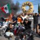 Manifestantes en Los Ángeles se congregan en una protesta con vehículos incendiados de fondo. Algunos portan la bandera de México mientras posan frente al fuego, en un acto de desafío que refleja el debate sobre la quema de símbolos nacionales y la libertad de expresión.