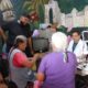 Dos mujeres adultas mayores reciben atención médica especializada en una jornada de salud visual en Huaquechula. Un médico y personal del Ayuntamiento acompañan la valoración en un espacio comunitario decorado con murales.