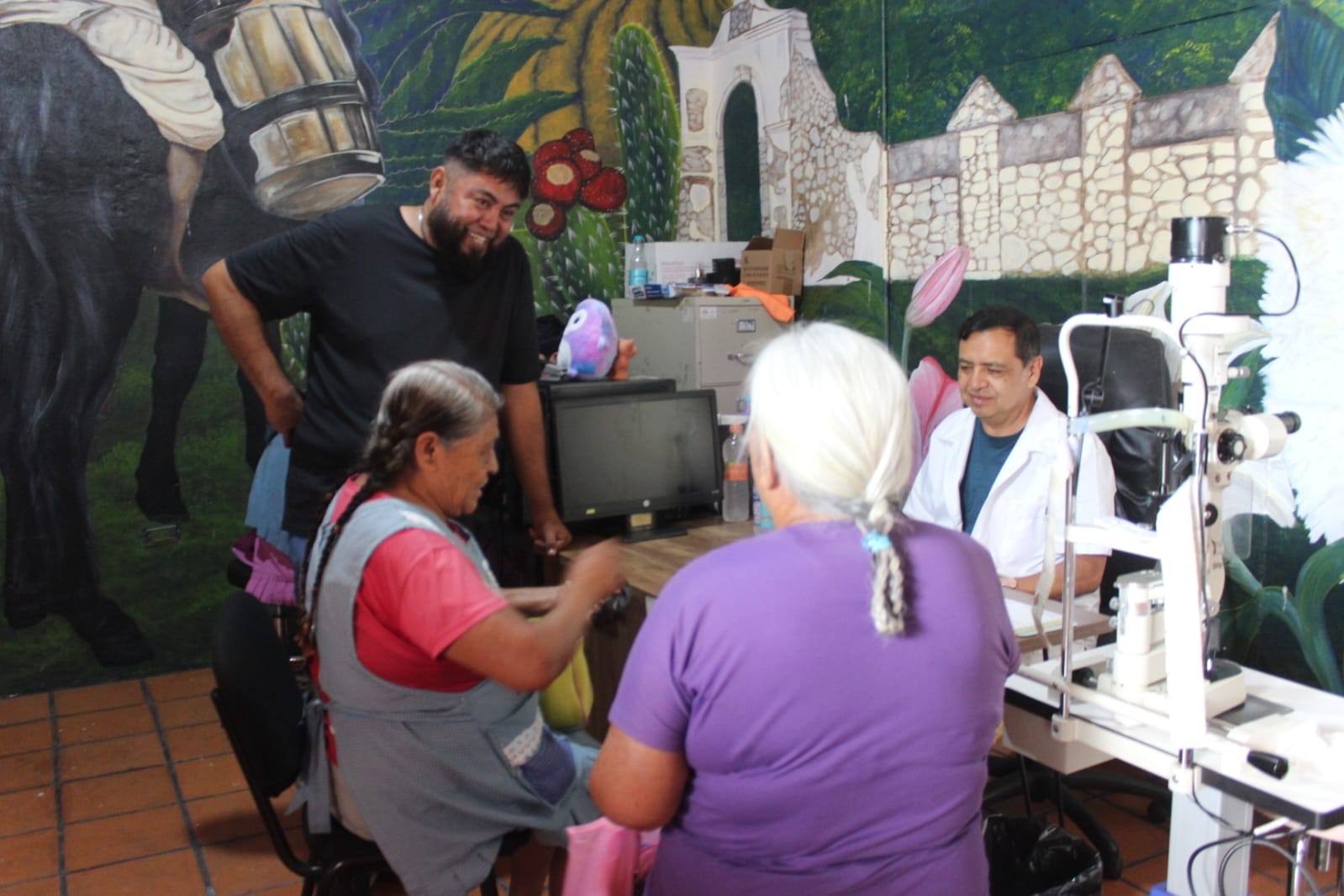 Dos mujeres adultas mayores reciben atención médica especializada en una jornada de salud visual en Huaquechula. Un médico y personal del Ayuntamiento acompañan la valoración en un espacio comunitario decorado con murales.