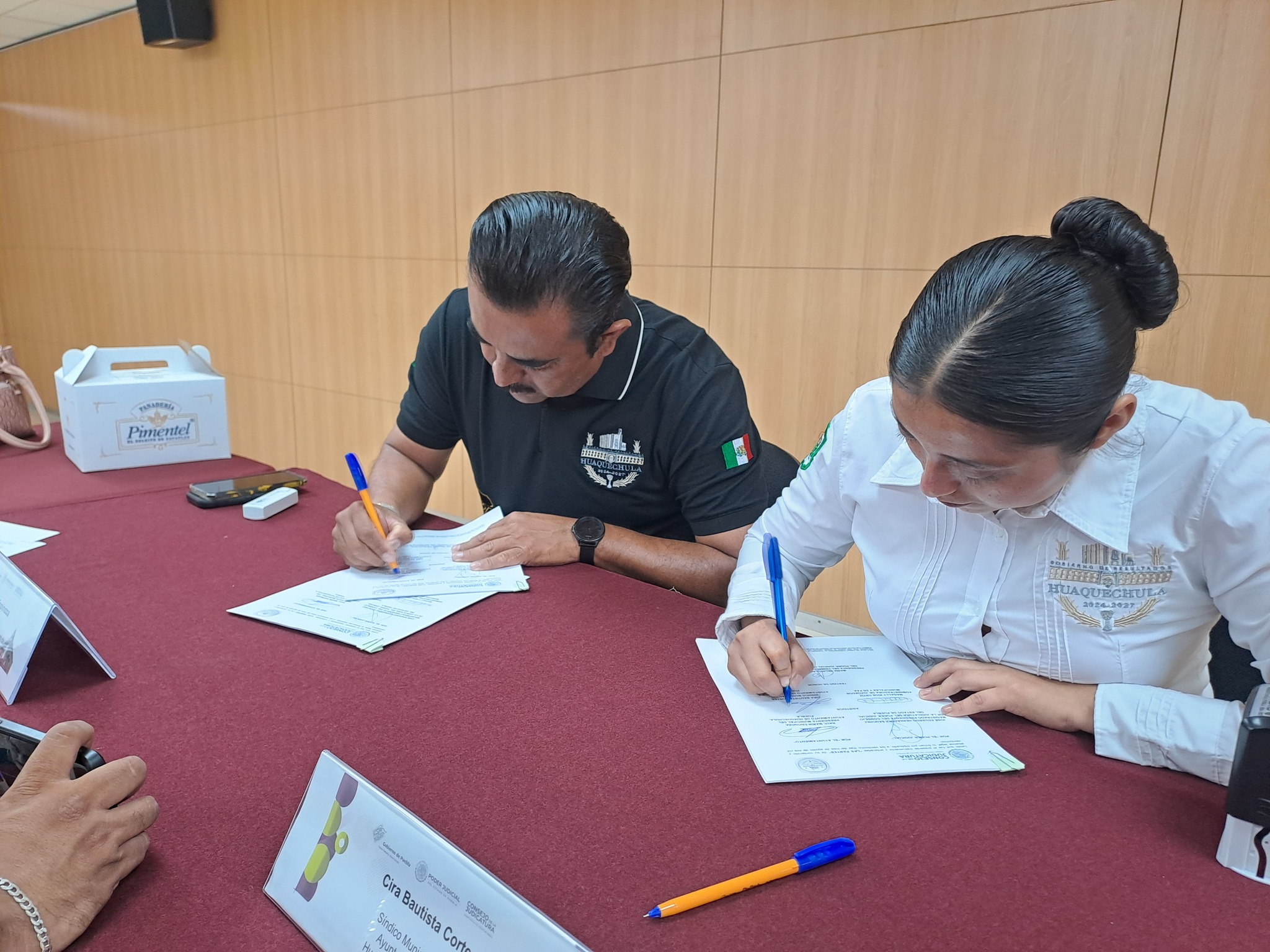 El presidente municipal de Huaquechula, Raúl Marín, y la Síndico Municipal firman un convenio de colaboración con el Poder Judicial del Estado de Puebla, en una mesa con documentos oficiales.