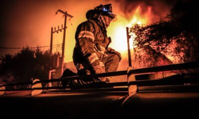 Bombero mexicano con uniforme completo y casco iluminado, arrodillado sobre una estructura frente a un incendio de gran magnitud durante la noche