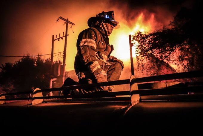 Bombero mexicano con uniforme completo y casco iluminado, arrodillado sobre una estructura frente a un incendio de gran magnitud durante la noche