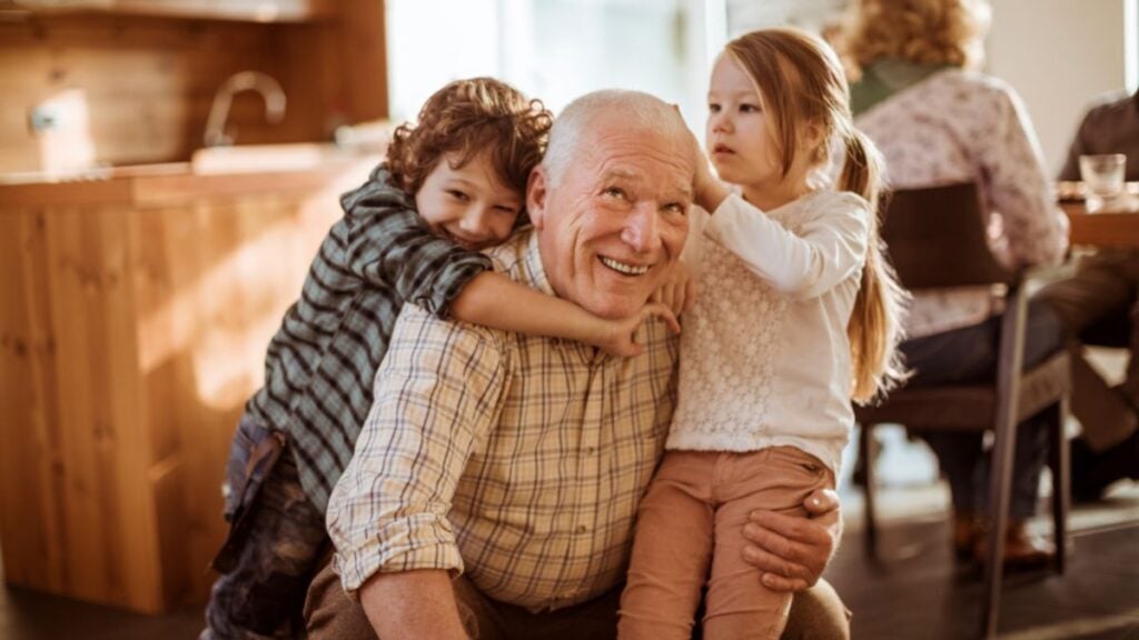 Un abuelo sonriente abraza a sus dos nietos pequeños en un ambiente familiar, reflejando unión, cariño y la importancia de los abuelos en la vida cotidiana.