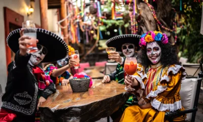 Personas con rostros pintados de calaveras y vestimenta tradicional mexicana celebran el Día de Muertos con bebidas típicas en una reunión comunitaria.