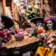 Personas con rostros pintados de calaveras y vestimenta tradicional mexicana celebran el Día de Muertos con bebidas típicas en una reunión comunitaria.