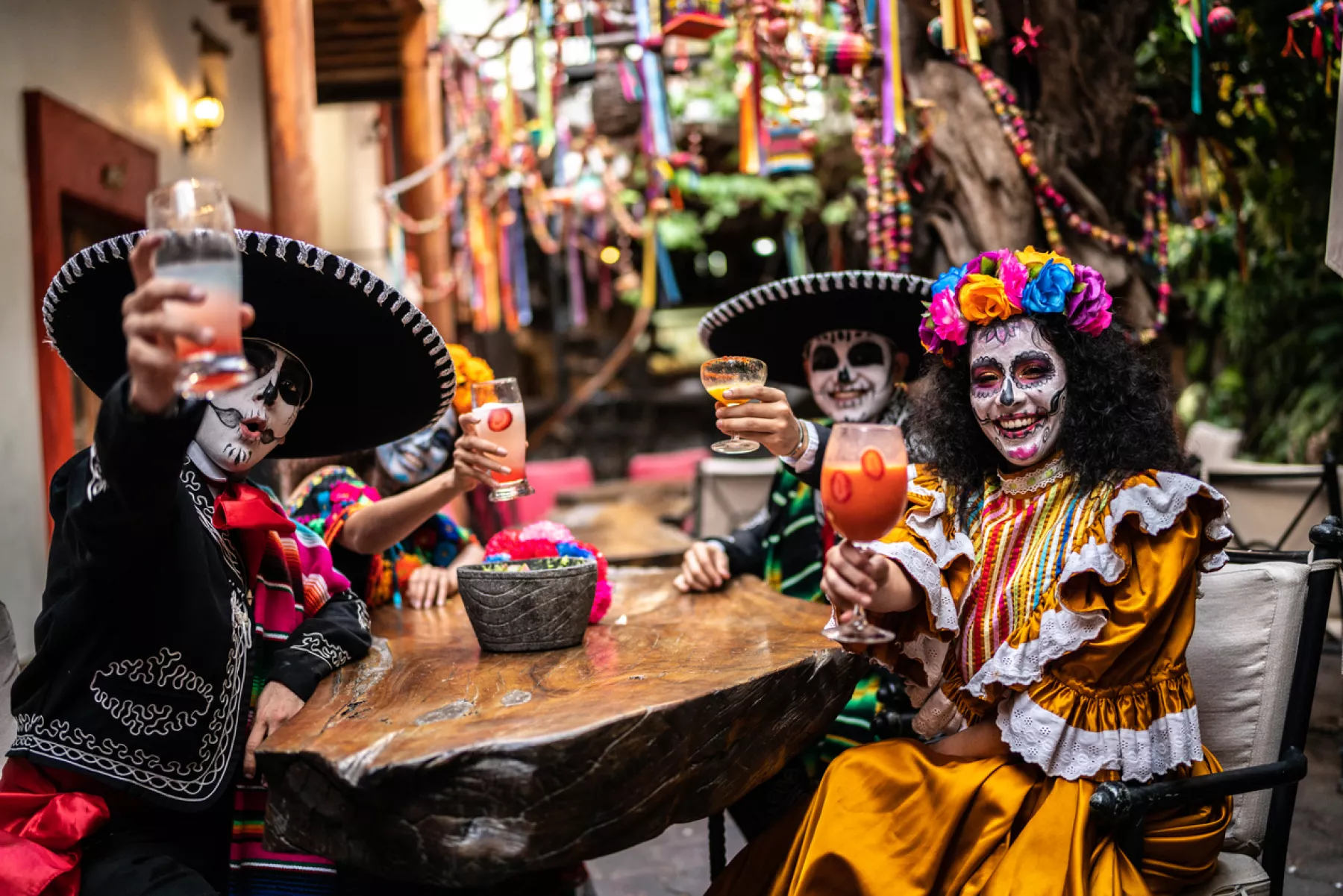 Personas con rostros pintados de calaveras y vestimenta tradicional mexicana celebran el Día de Muertos con bebidas típicas en una reunión comunitaria.