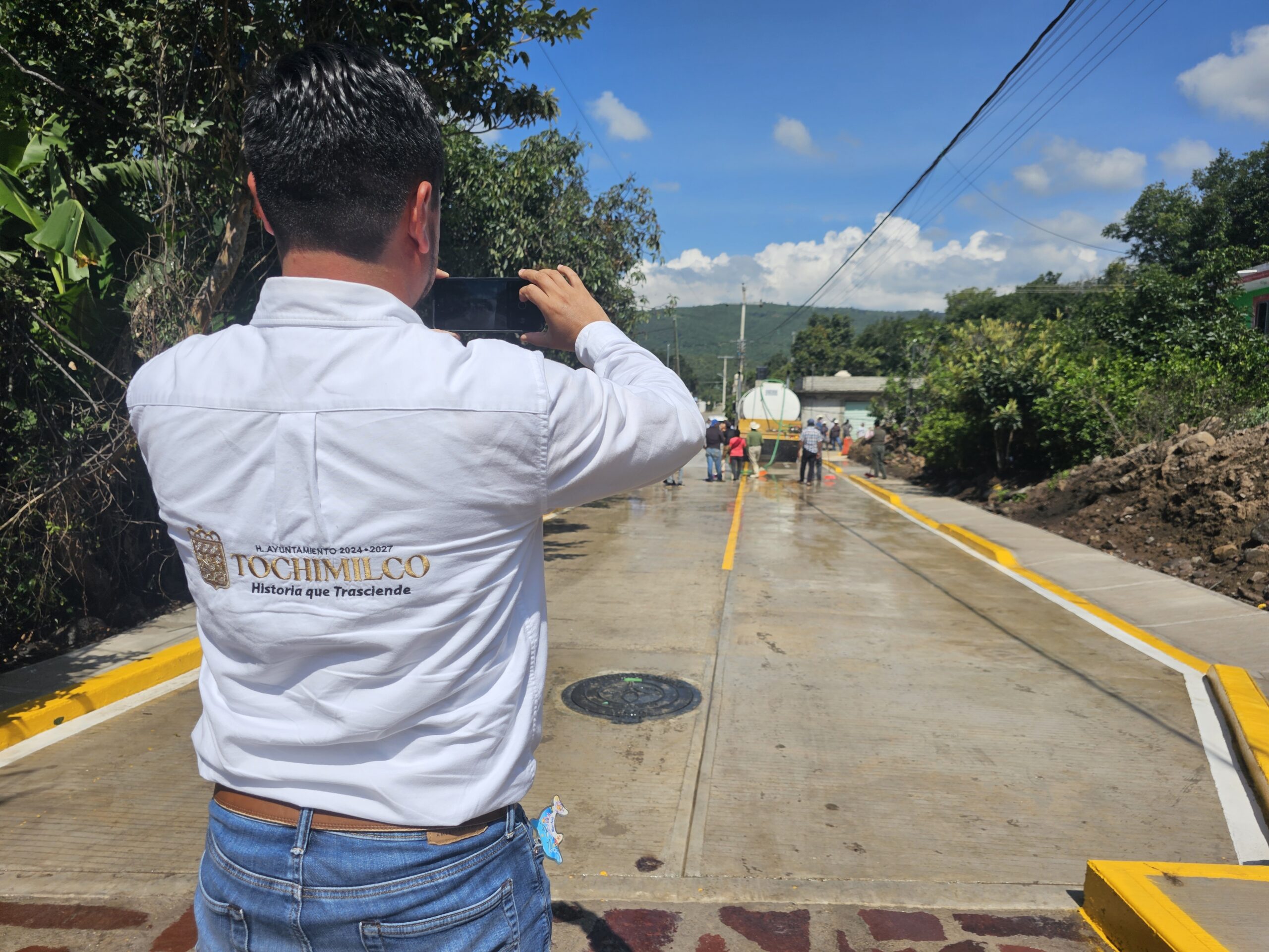 Persona con camisa institucional del Ayuntamiento de Tochimilco fotografía la nueva calle Miguel Hidalgo, recién pavimentada con concreto hidráulico, mientras vecinos observan la inauguración de la obra.