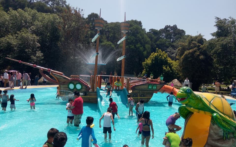 Niños y adultos disfrutan de un área acuática infantil en un balneario. Se observa una alberca con toboganes, un barco pirata decorativo y una resbaladilla en forma de tortuga, en un ambiente familiar y recreativo.