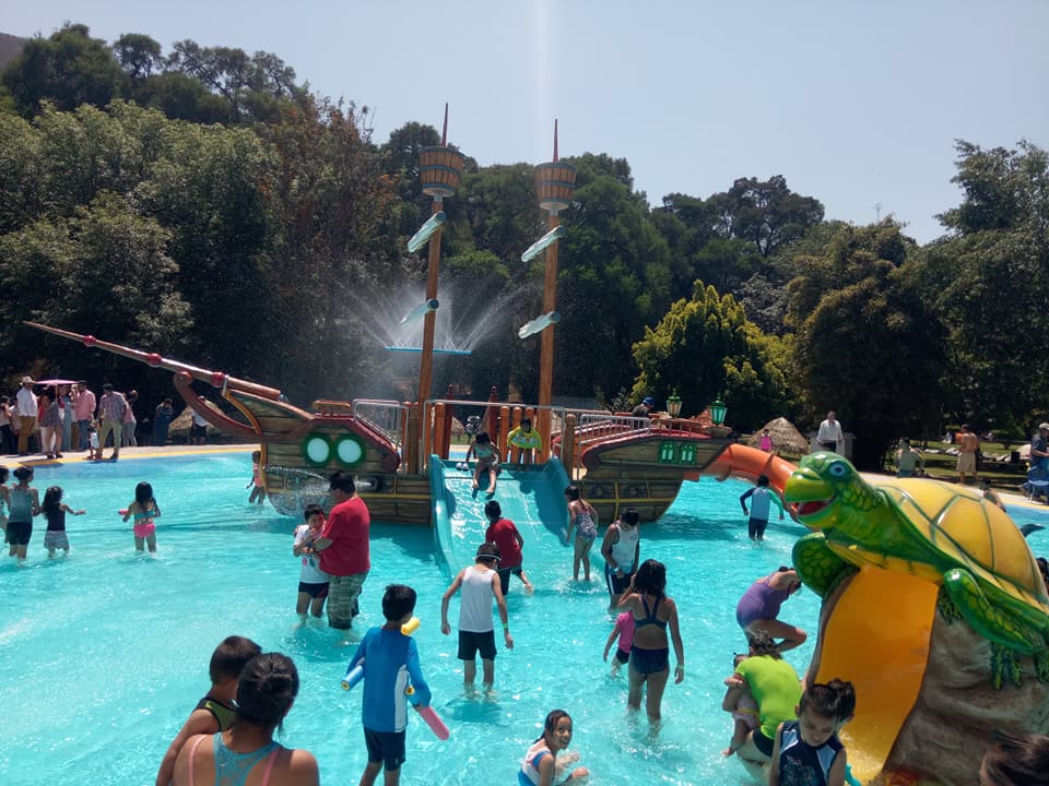 Niños y adultos disfrutan de un área acuática infantil en un balneario. Se observa una alberca con toboganes, un barco pirata decorativo y una resbaladilla en forma de tortuga, en un ambiente familiar y recreativo.