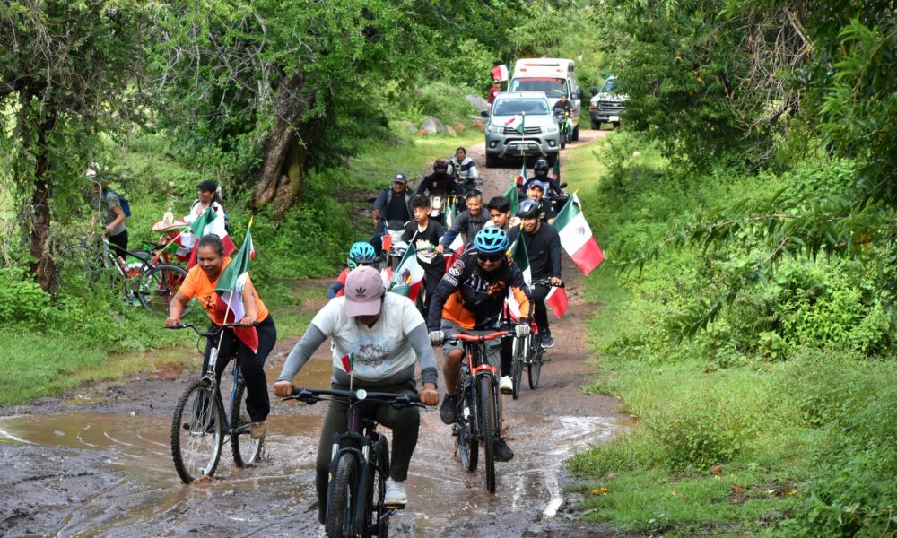 Grupo de ciclistas avanza por un camino rural con banderas de México, durante la 3ª Rodada Familiar en Huaquechula, organizada en el marco de las Fiestas Patrias.