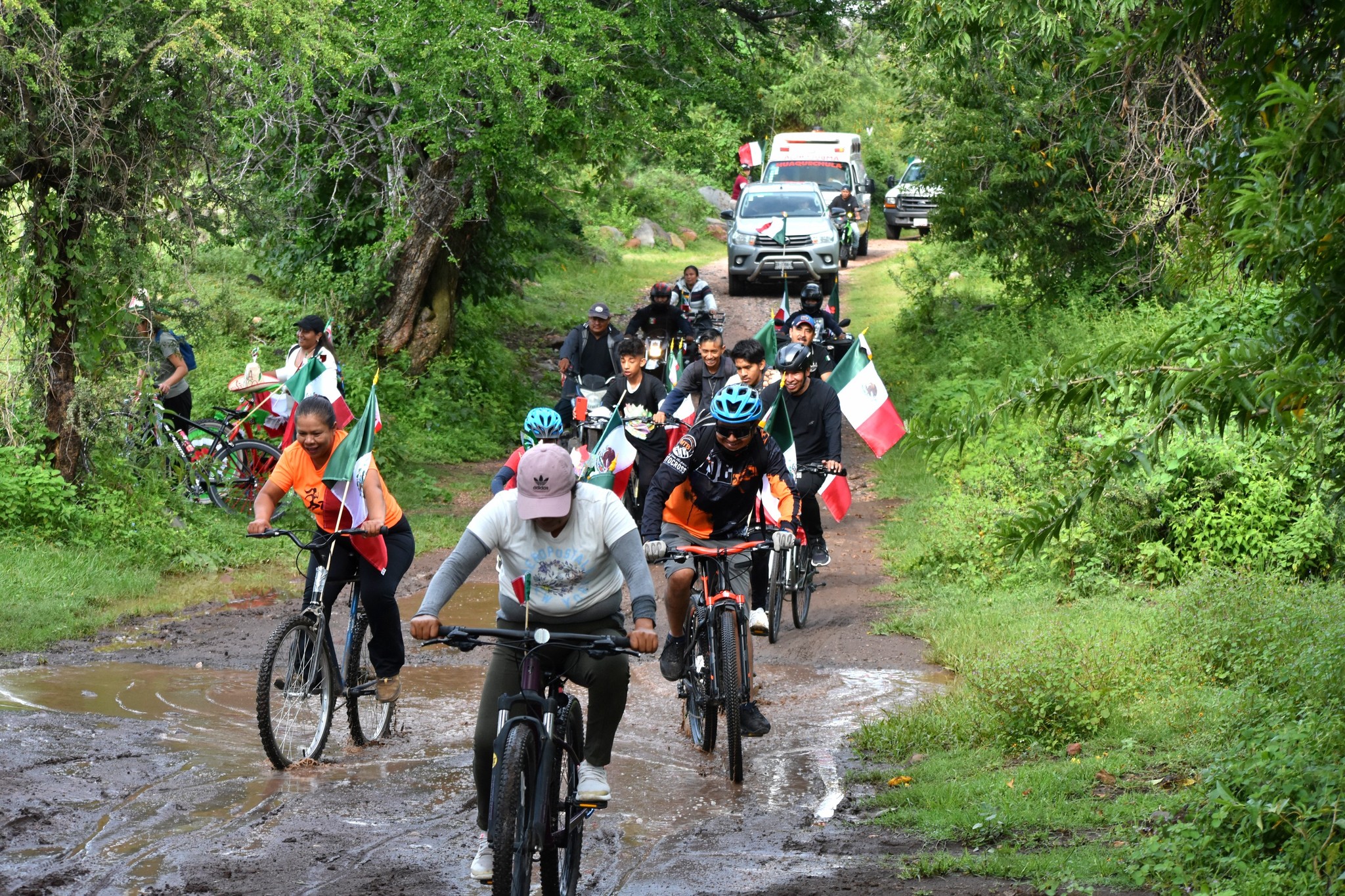 Grupo de ciclistas avanza por un camino rural con banderas de México, durante la 3ª Rodada Familiar en Huaquechula, organizada en el marco de las Fiestas Patrias.