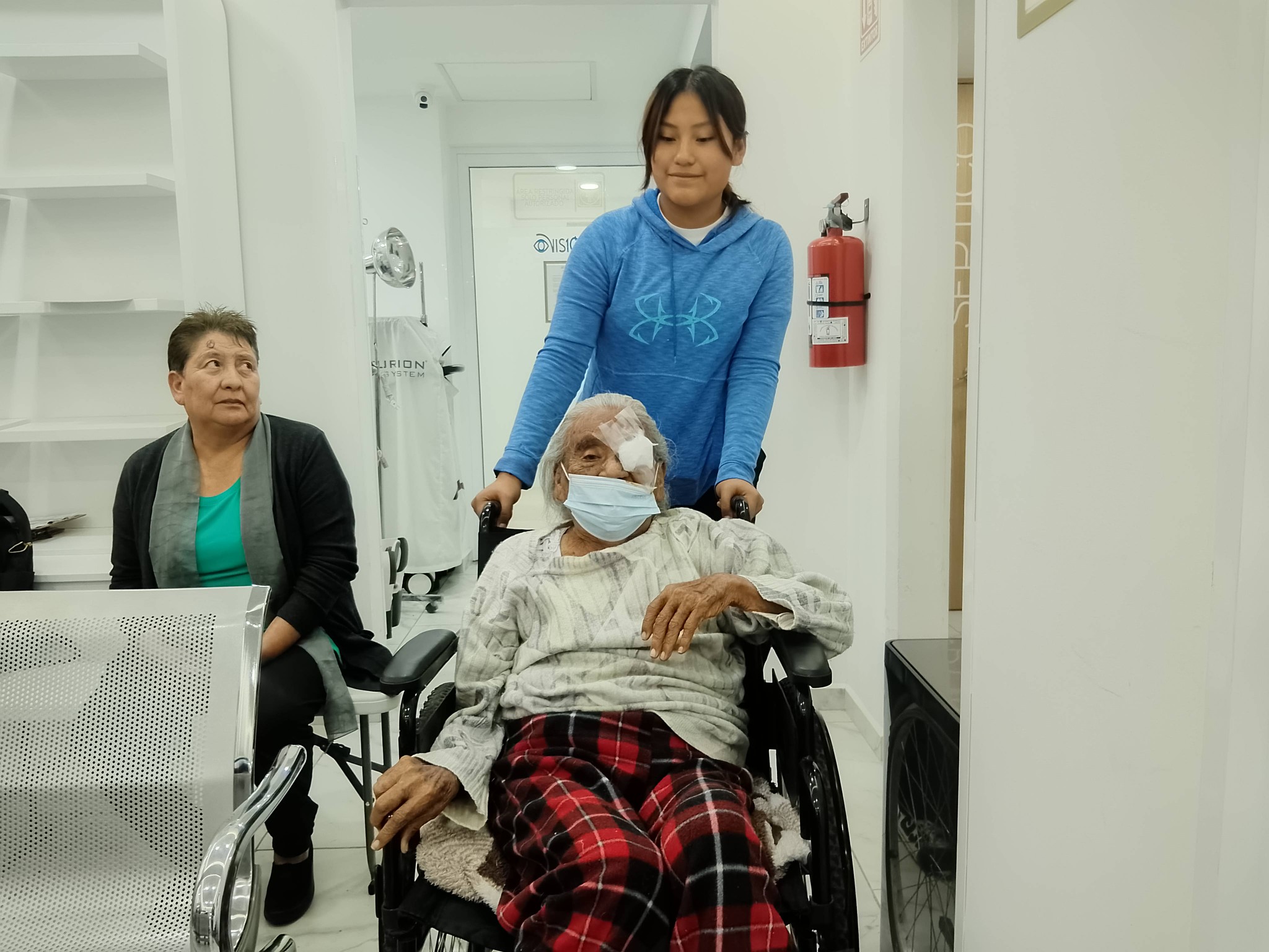 Adulto mayor en silla de ruedas con un parche en el ojo tras una cirugía de cataratas, acompañado por familiares en la sala de una clínica.