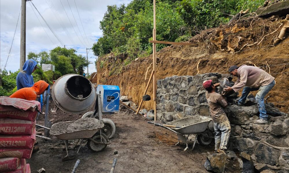 Trabajadores de la construcción colocan piedra y cemento en un muro de contención junto a una calle en obra, mientras se utiliza una mezcladora de concreto. La escena muestra el avance de la pavimentación en Santiago Tochimizolco.