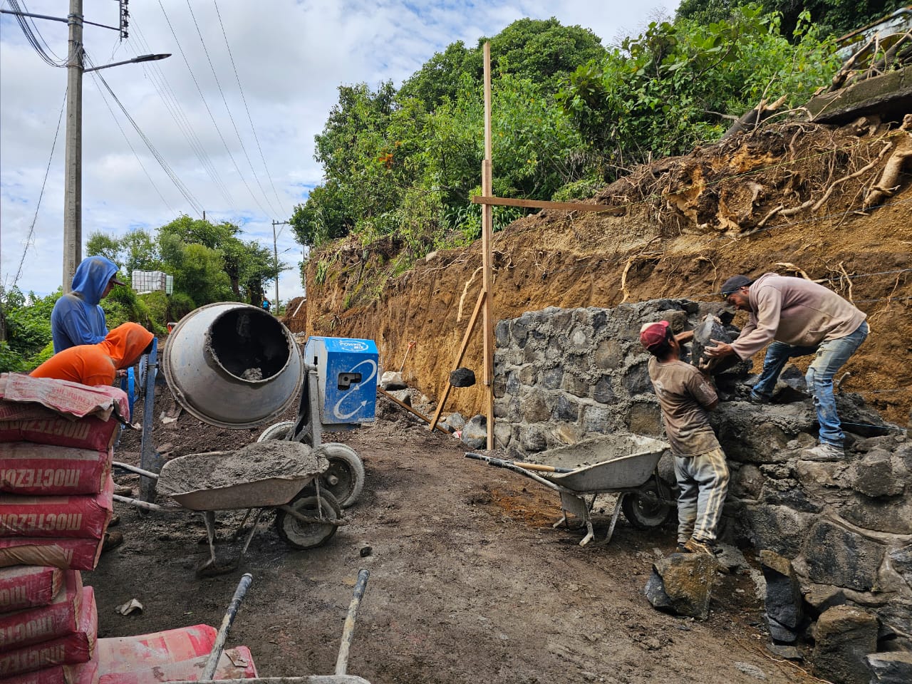 Trabajadores de la construcción colocan piedra y cemento en un muro de contención junto a una calle en obra, mientras se utiliza una mezcladora de concreto. La escena muestra el avance de la pavimentación en Santiago Tochimizolco.