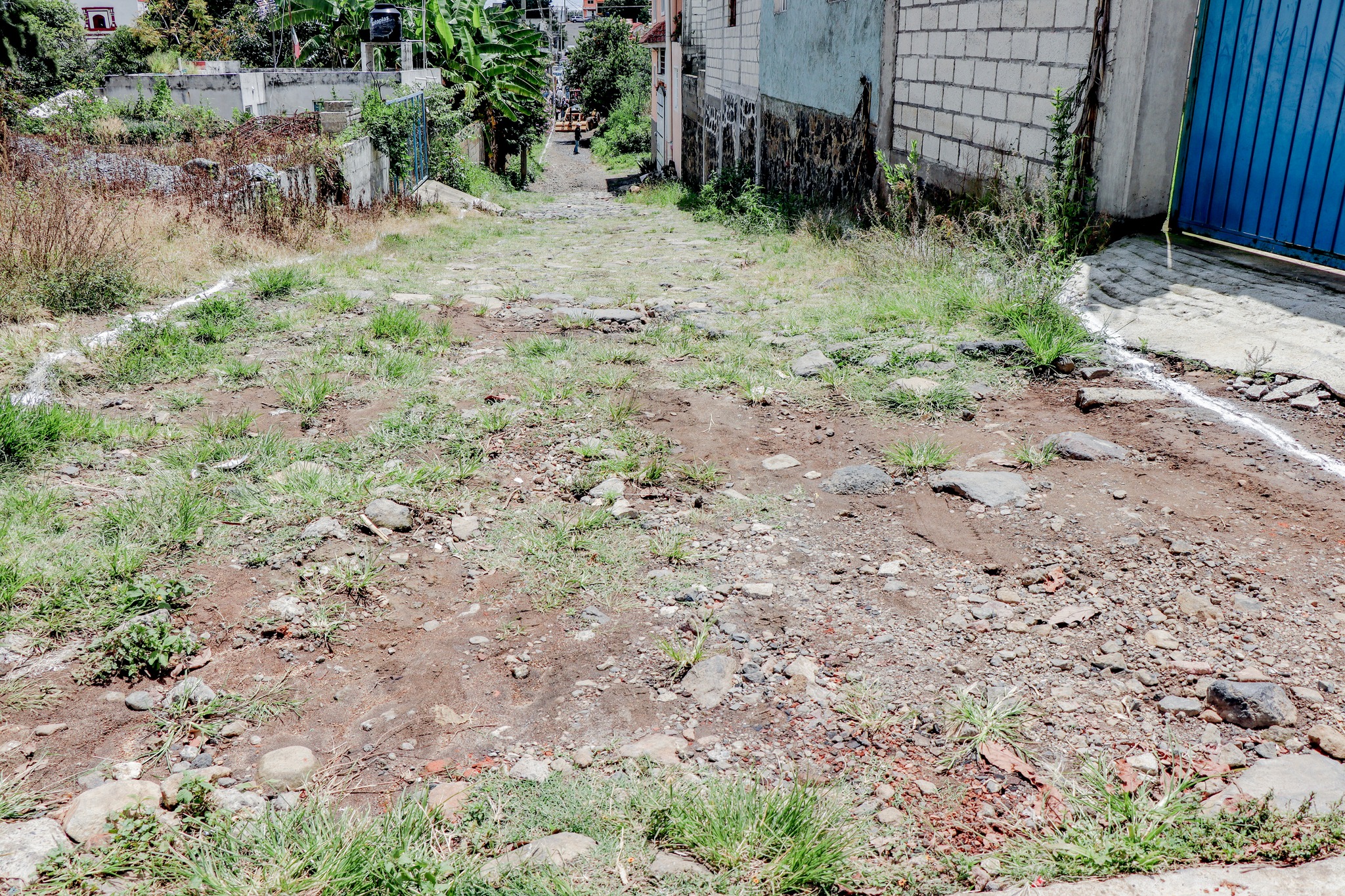Vista de una calle sin pavimentar en San Lucas Tulcingo, con superficie de tierra y piedras, rodeada de viviendas y vegetación. Se observan marcas de cal en los bordes que delimitan la zona destinada a obra pública.