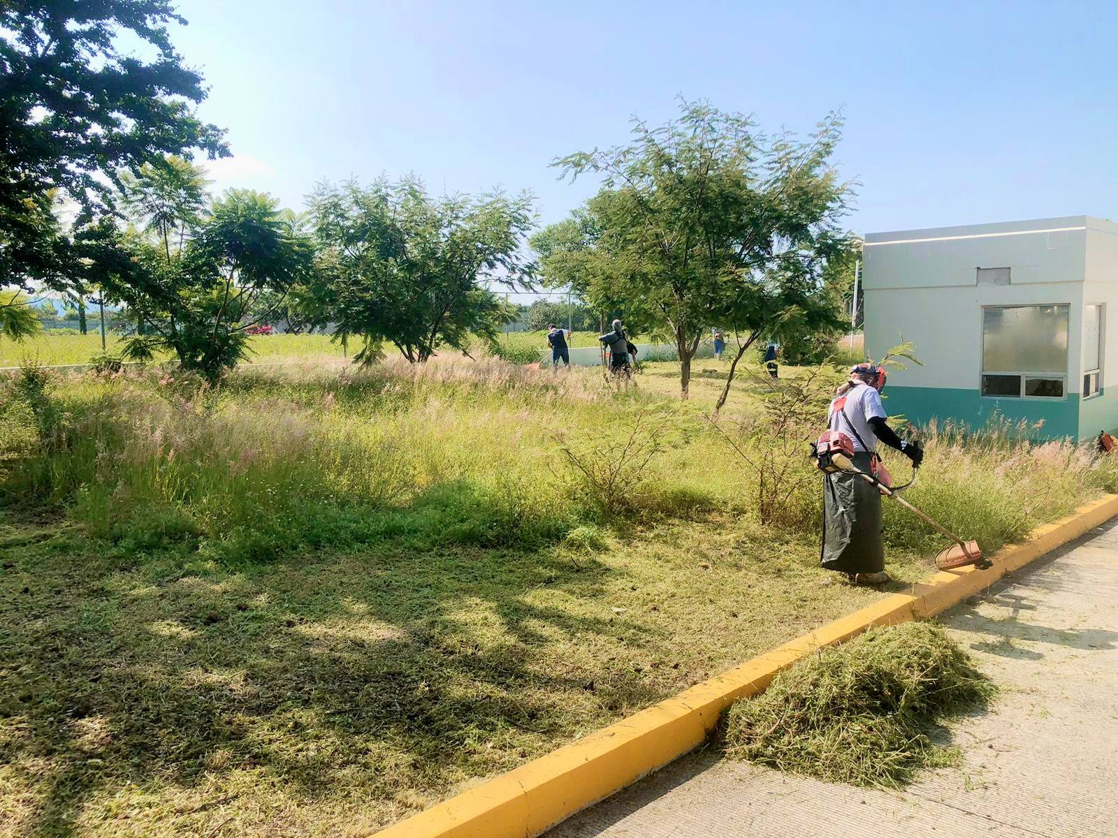 Personas realizan labores de limpieza y deshierbe en áreas verdes frente a un centro de salud en Cacaloxúchitl. Un trabajador con equipo de protección corta el pasto con desbrozadora, mientras otros participantes colaboran en la faena comunitaria.