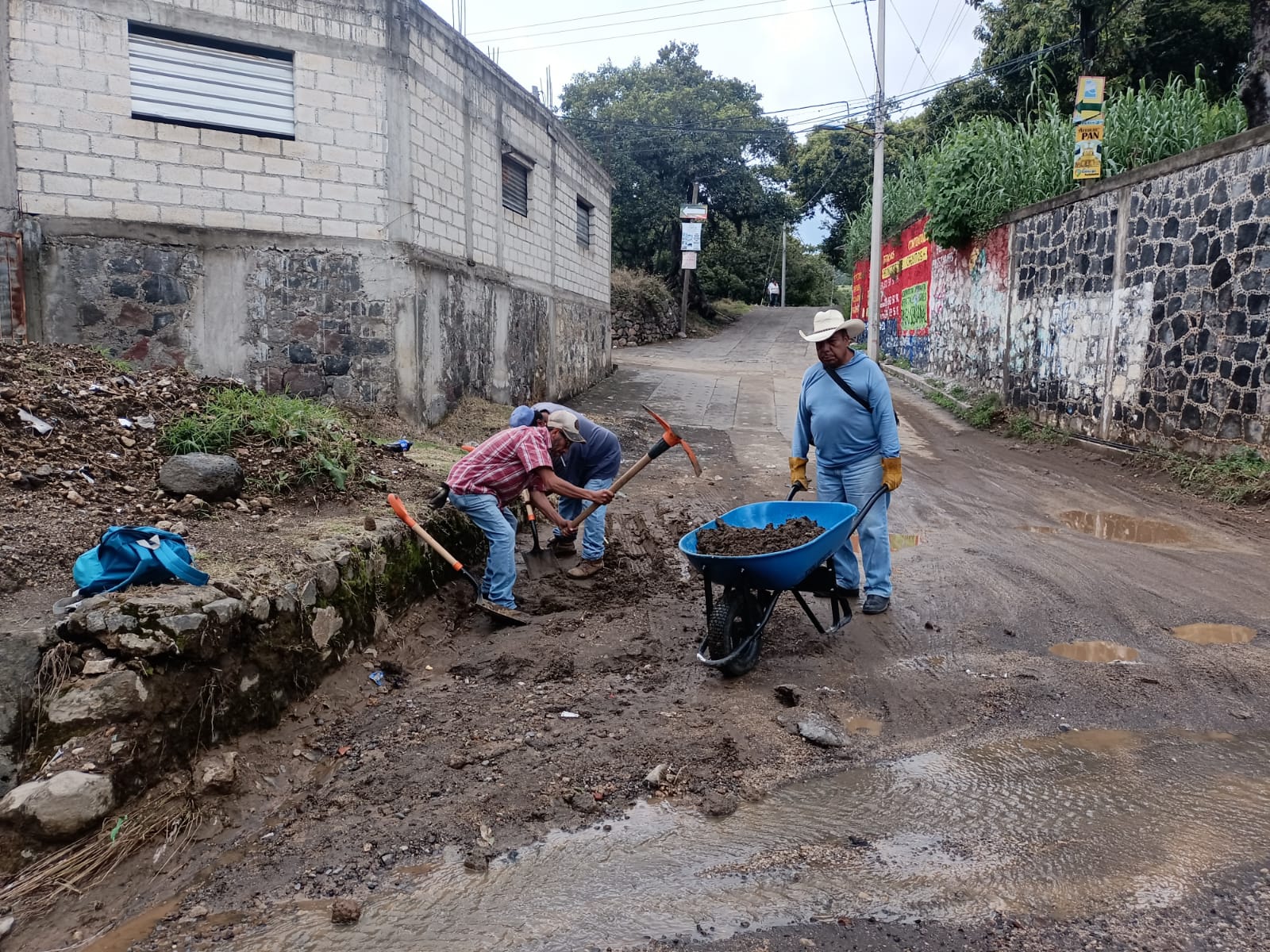 Personal de Servicios Públicos del Ayuntamiento de Tochimilco realizando limpieza y retiro de escombros en vialidades tras lluvias intensas, con herramientas y carretilla en una calle afectada.