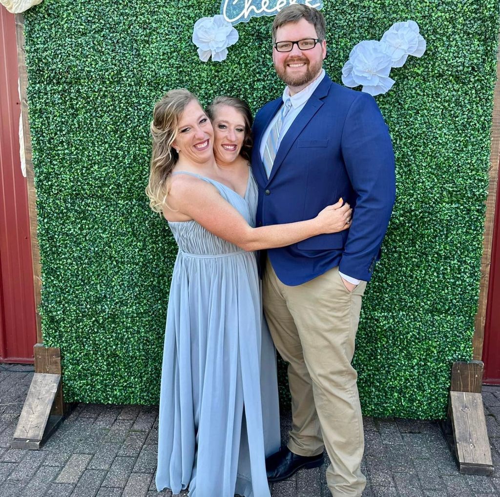 Abby y Brittany Hensel, gemelas siamesas, posan sonrientes junto a Josh Bowling, esposo de Abby, frente a un fondo decorativo verde durante un evento familiar.