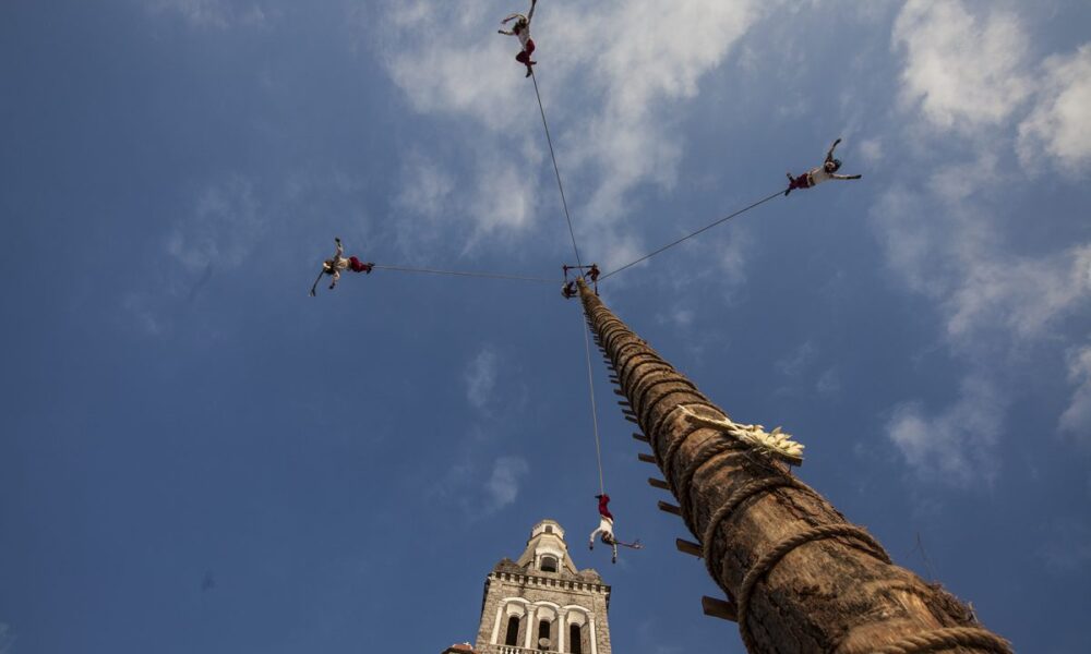 Fotografía tomada desde el suelo hacia el cielo, mostrando a los Voladores de Cuetzalan descendiendo con cuerdas desde un alto mástil ceremonial frente a la iglesia, enmarcados por un cielo azul con nubes.