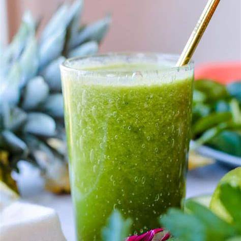 Vaso de jugo verde fresco con pajilla dorada, rodeado de frutas como piña y limón, sobre una mesa con ingredientes naturales.