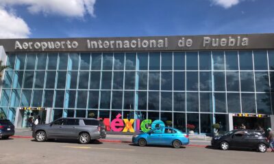 Fachada del Aeropuerto Internacional de Puebla “Hermanos Serdán”, con autos en el acceso principal y el letrero colorido de México al frente.