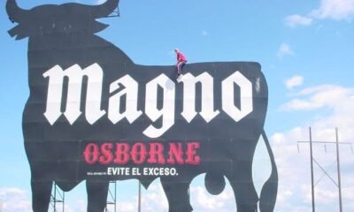 Persona subiendo al icónico anuncio metálico del toro de Magno Osborne, clásico emblema publicitario visible en carreteras.