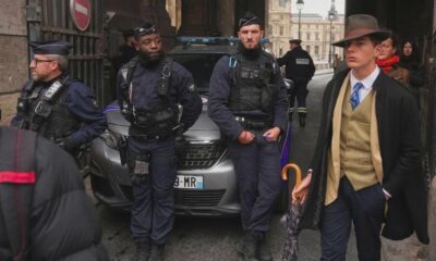 Detective elegante con sombrero y paraguas de pie junto a policías franceses en la entrada del Museo del Louvre.