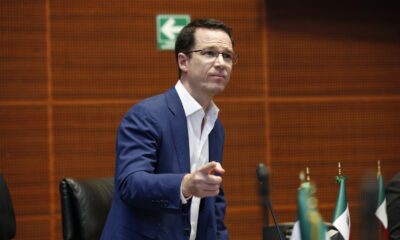 Persona con traje azul hablando frente a un micrófono y banderas de México durante una sesión política.
