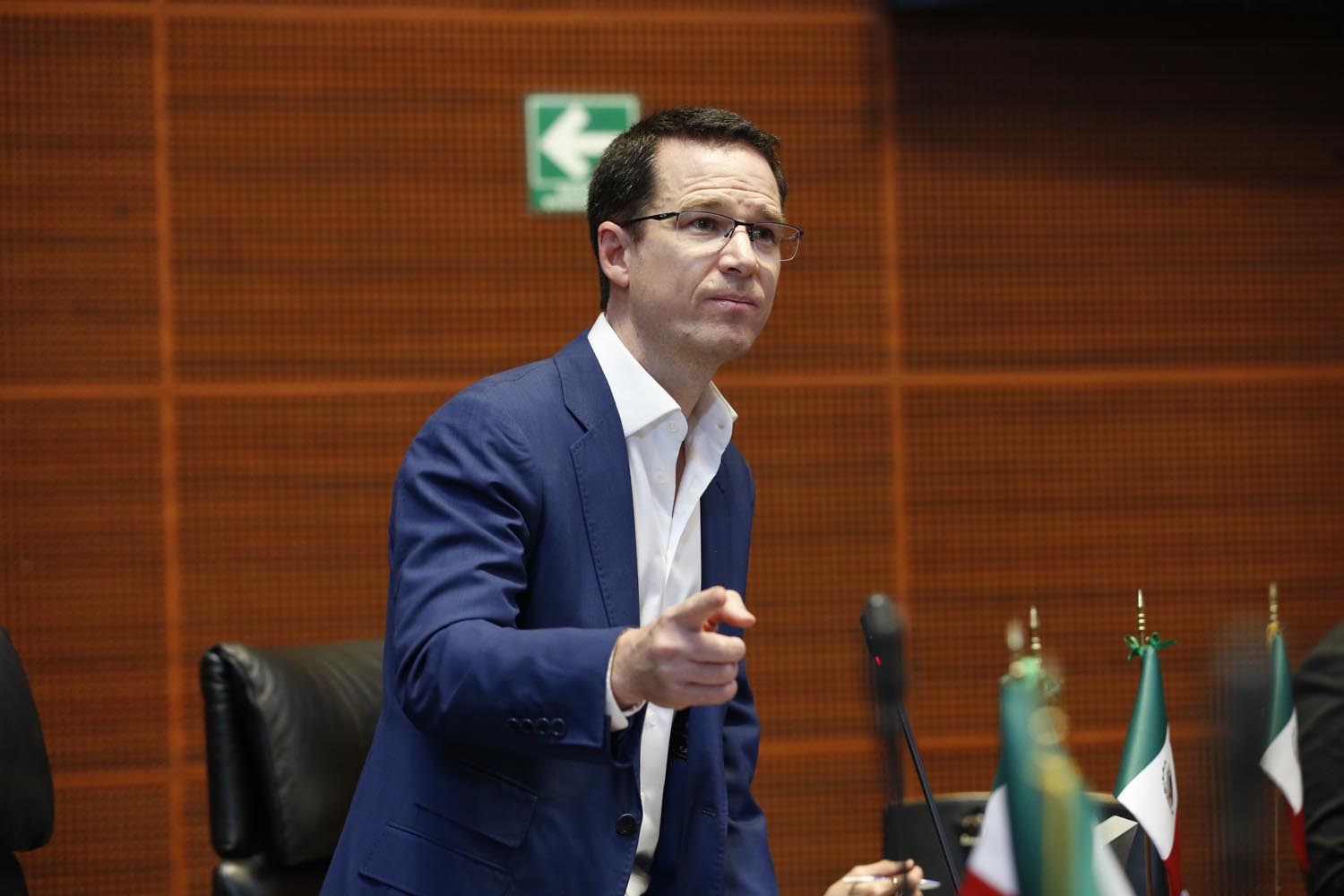 Persona con traje azul hablando frente a un micrófono y banderas de México durante una sesión política.