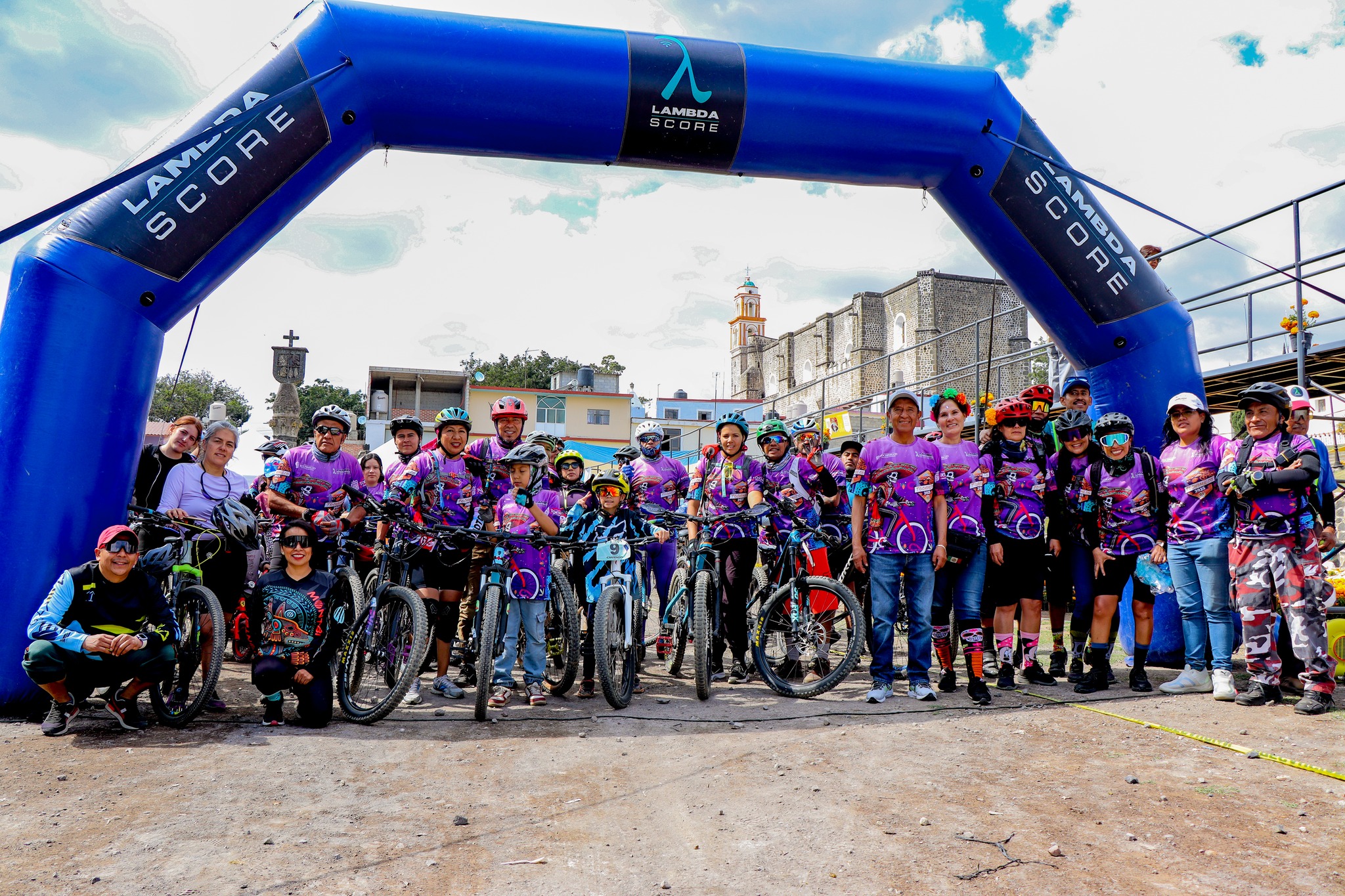 Participantes de la 5ta edición deportiva “Ánimas y Recuerdos” en Tochimilco, bajo el arco de salida, con bicicletas y vestimenta alusiva al Día de Muertos.