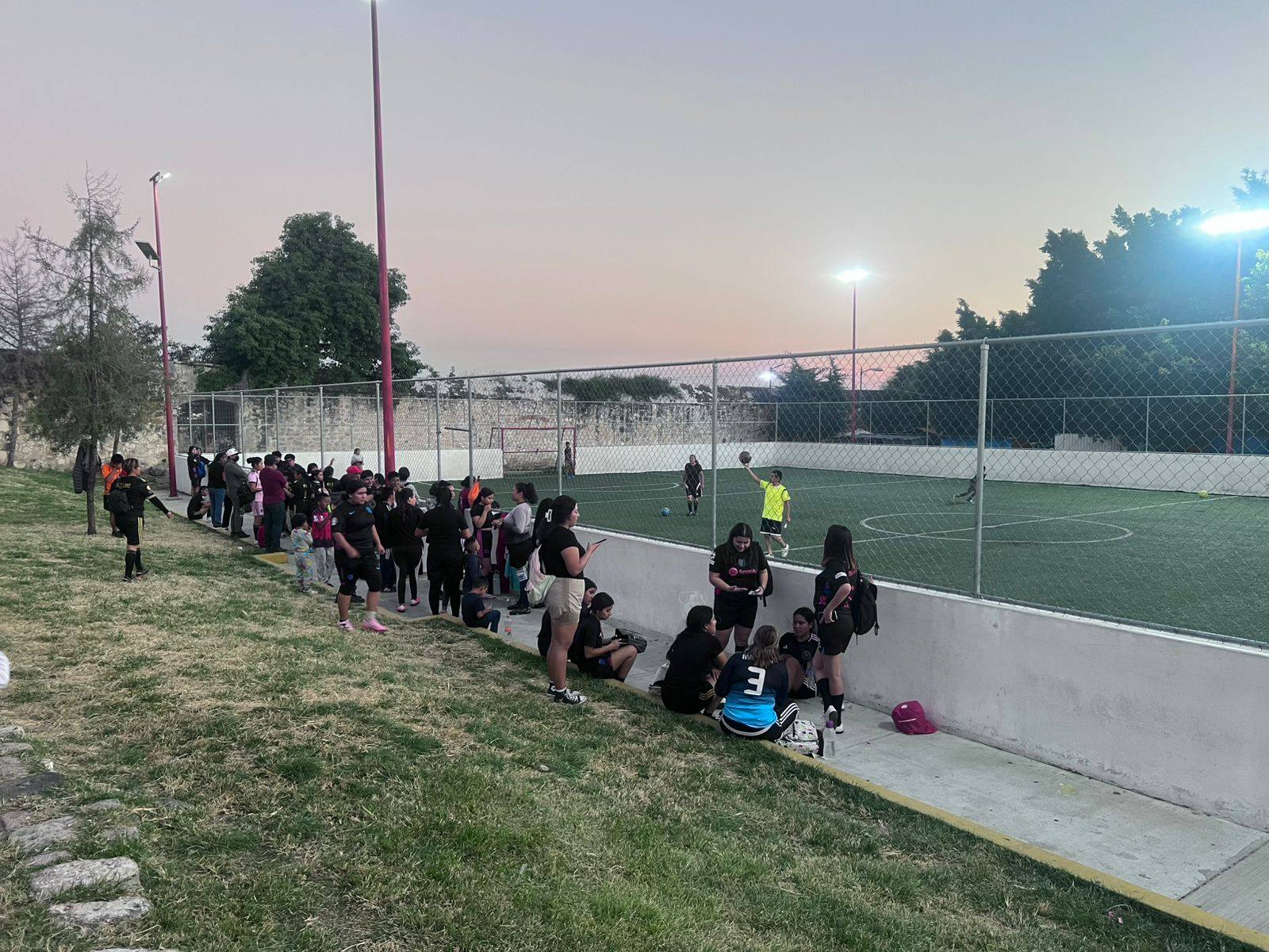 Personas reunidas alrededor de una cancha de fútbol rápido al atardecer durante partidos comunitarios en Huaquechula.