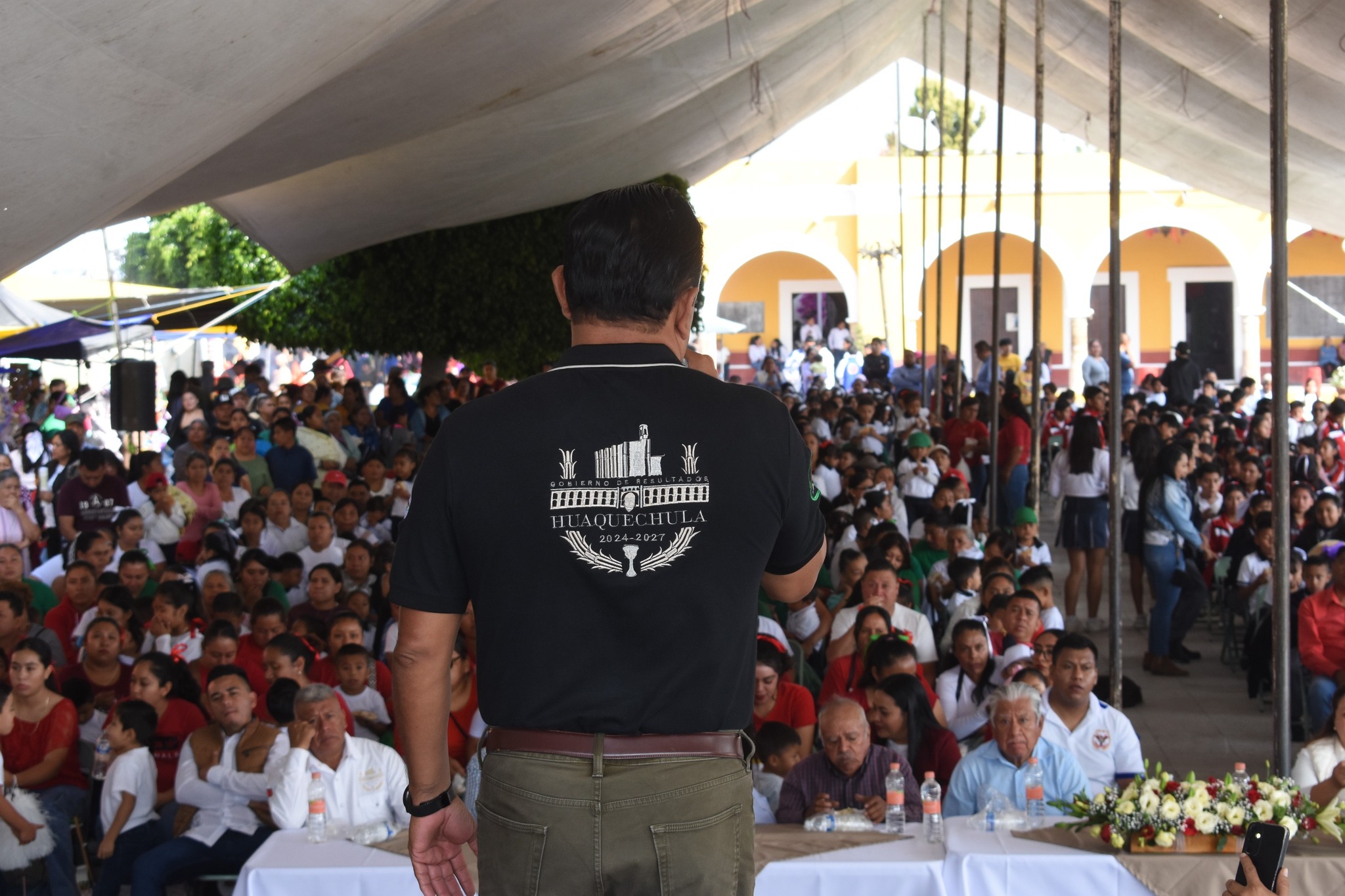 Presidente municipal de Huaquechula frente a un público numeroso bajo una carpa, durante la ceremonia cívica del 215 aniversario de la Revolución Mexicana.