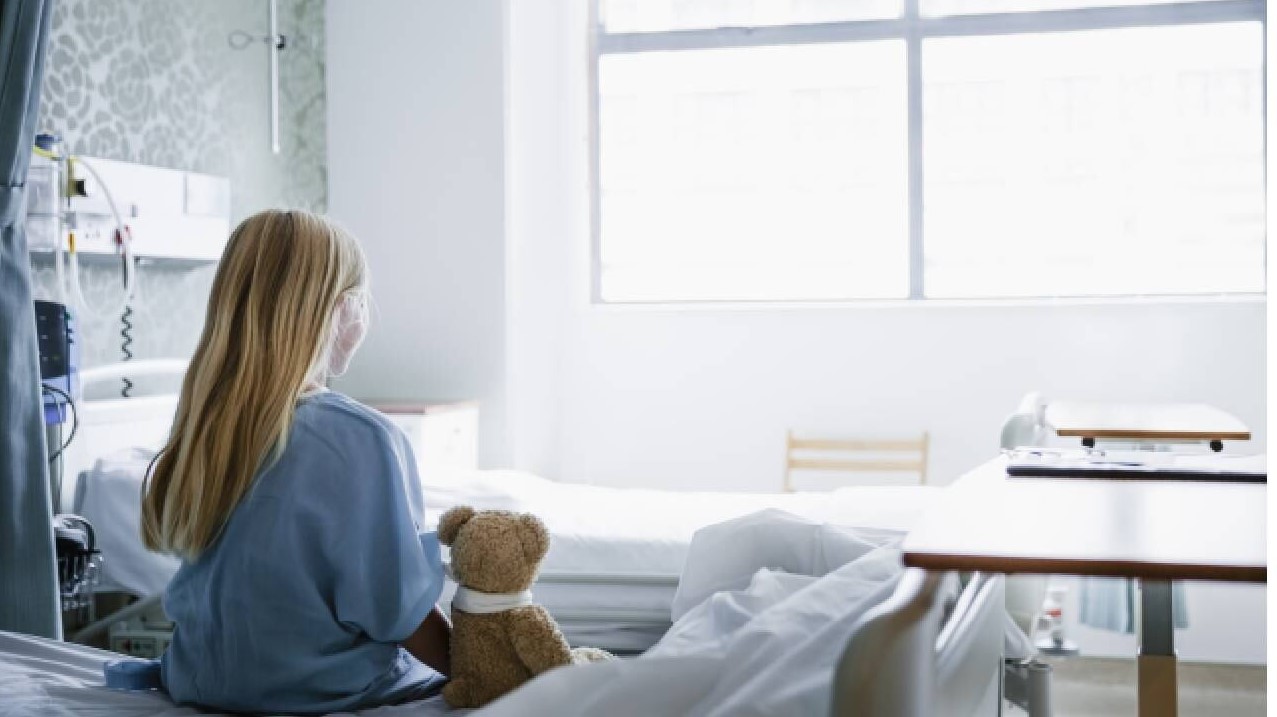 Niña sentada sobre una cama blanca en una habitación iluminada, mirando hacia una ventana. Imagen ilustrativa del caso de Olivia Farnsworth.