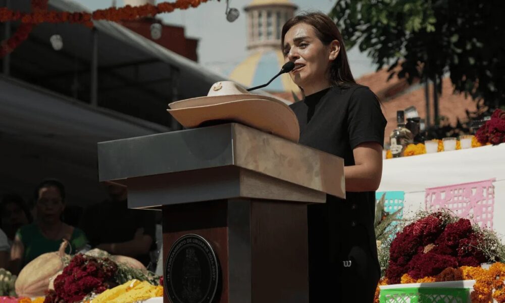 Mujer con expresión de tristeza ofrece un discurso público frente a un micrófono; en primer plano, un sombrero de ala ancha sobre el podio.