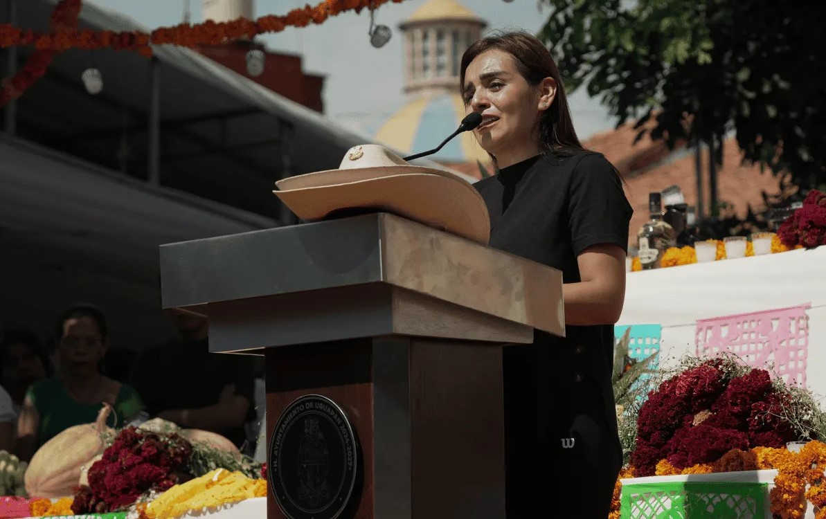 Mujer con expresión de tristeza ofrece un discurso público frente a un micrófono; en primer plano, un sombrero de ala ancha sobre el podio.