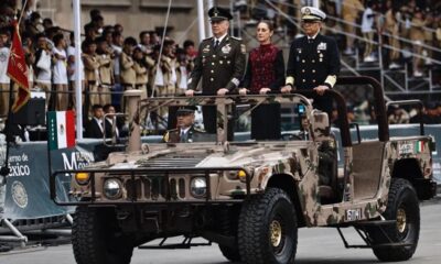 Vehículo militar con autoridades nacionales participando en desfile público frente a asistentes y banderas de México.
