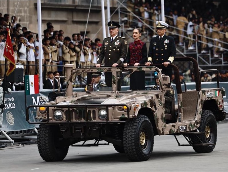 Vehículo militar con autoridades nacionales participando en desfile público frente a asistentes y banderas de México.