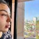 Mujer joven con lesiones visibles en rostro y frente observa por la ventana hacia una ciudad; representa denuncia de violencia policial y debate sobre justicia y derechos humanos.