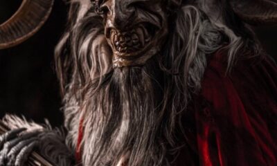 Personaje caracterizado como Krampus, criatura con cuernos y máscara oscura, vestido con pieles y capa roja, sosteniendo un bastón, en un ambiente oscuro y frío.