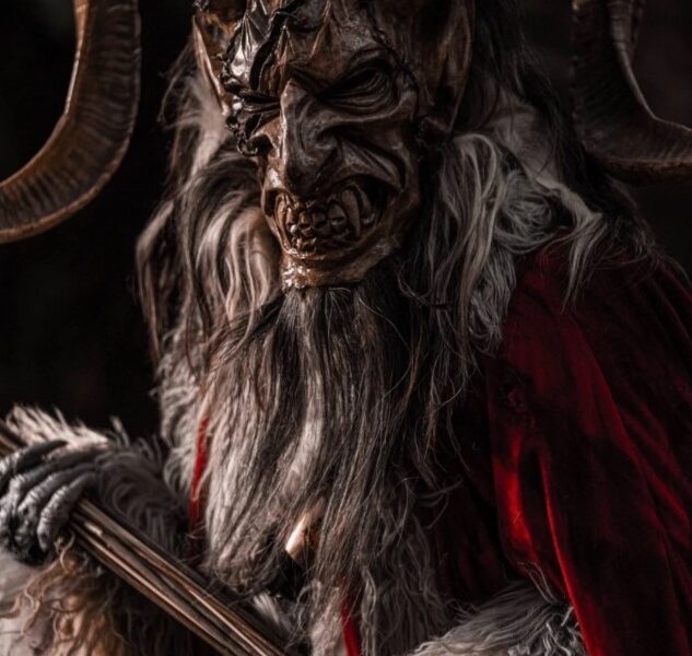 Personaje caracterizado como Krampus, criatura con cuernos y máscara oscura, vestido con pieles y capa roja, sosteniendo un bastón, en un ambiente oscuro y frío.