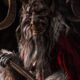 Personaje caracterizado como Krampus, criatura con cuernos y máscara oscura, vestido con pieles y capa roja, sosteniendo un bastón, en un ambiente oscuro y frío.
