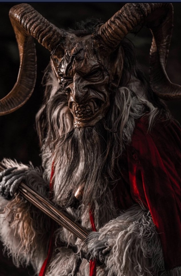 Personaje caracterizado como Krampus, criatura con cuernos y máscara oscura, vestido con pieles y capa roja, sosteniendo un bastón, en un ambiente oscuro y frío.