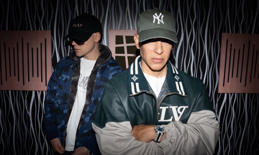 Bizarrap y Daddy Yankee posan en un estudio de grabación, vestidos con chamarras deportivas y gorras, antes del estreno de su nueva colaboración musical.