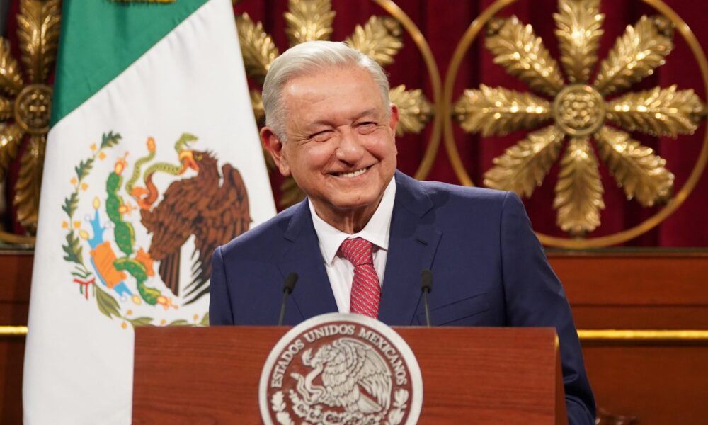 Expresidente de México sonriendo detrás de un podio oficial, con la bandera nacional al fondo.