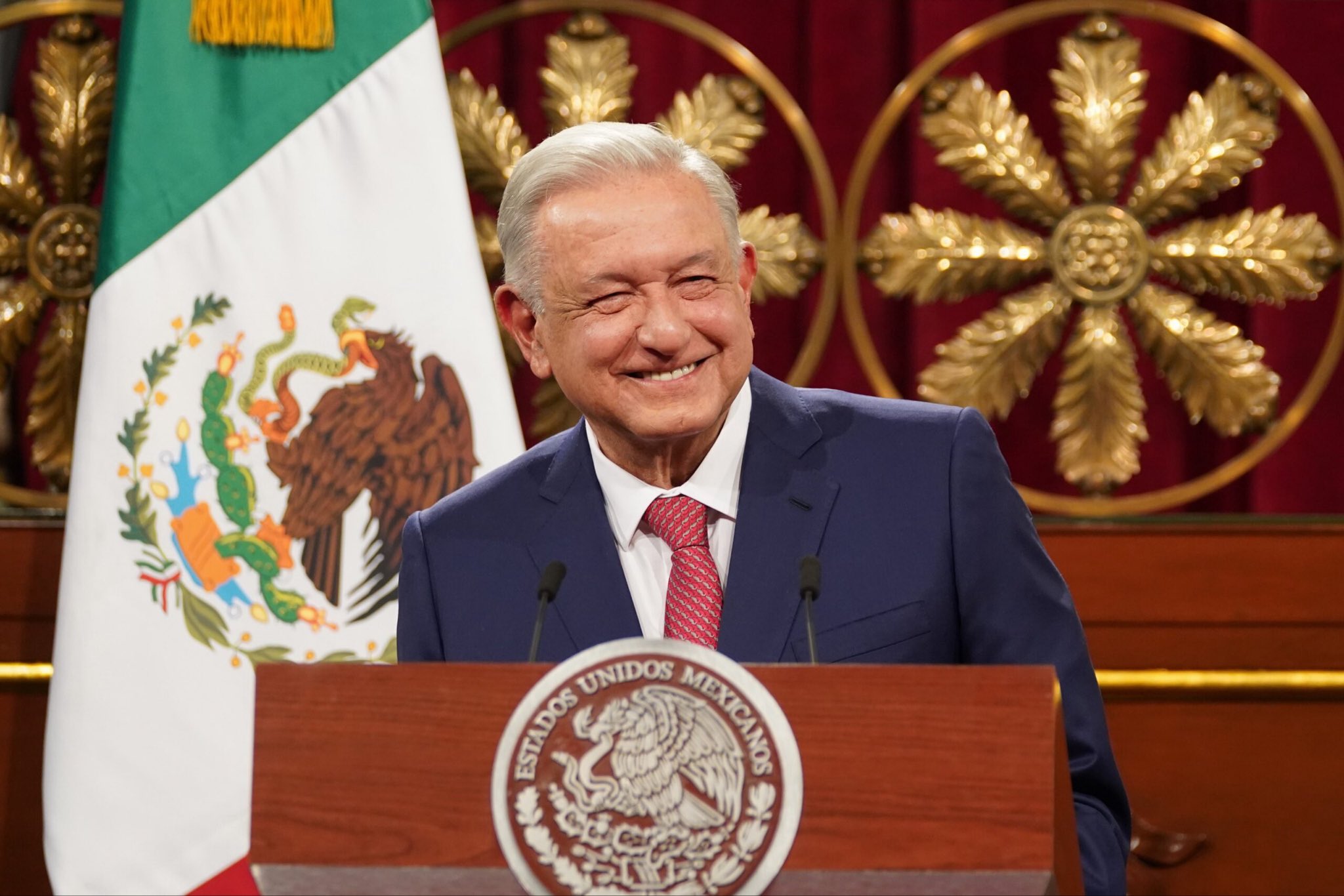 Expresidente de México sonriendo detrás de un podio oficial, con la bandera nacional al fondo.