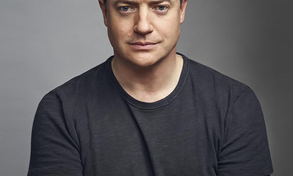 Retrato frontal de Brendan Fraser con camisa oscura, brazos cruzados y expresión seria, posando sobre un fondo gris neutro.