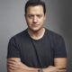 Retrato frontal de Brendan Fraser con camisa oscura, brazos cruzados y expresión seria, posando sobre un fondo gris neutro.