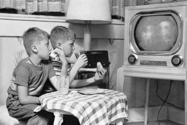 Dos niños observando atentos un televisor antiguo en blanco y negro mientras comen, representando la nostalgia y el impacto histórico de la televisión en la vida cotidiana.