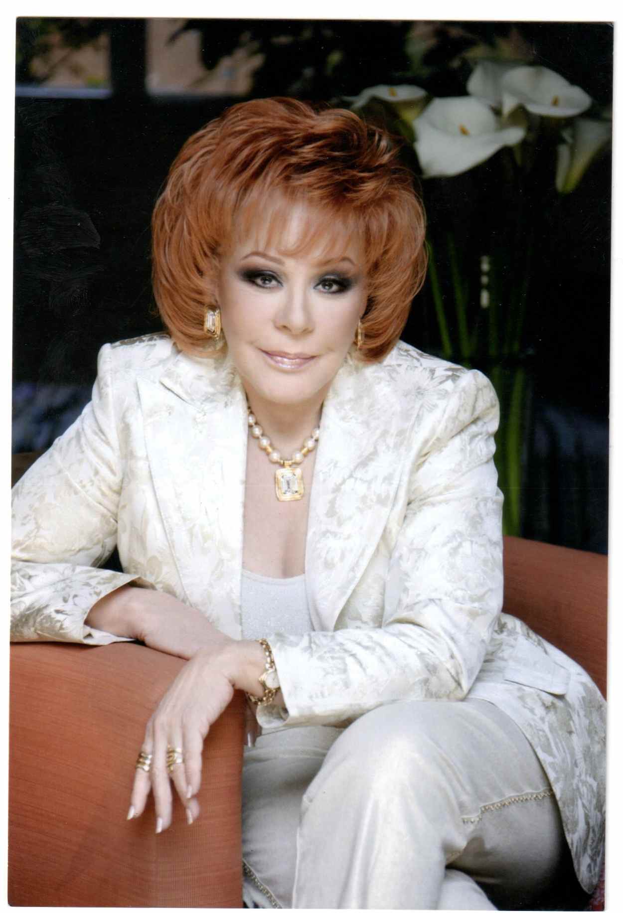 Retrato formal de una figura femenina del entretenimiento mexicano, con atuendo elegante claro, sentada en un sillón dentro de un ambiente interior.